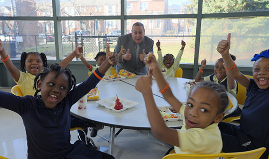 children at a lunch table reaching out with their arms in the air and thumbs up